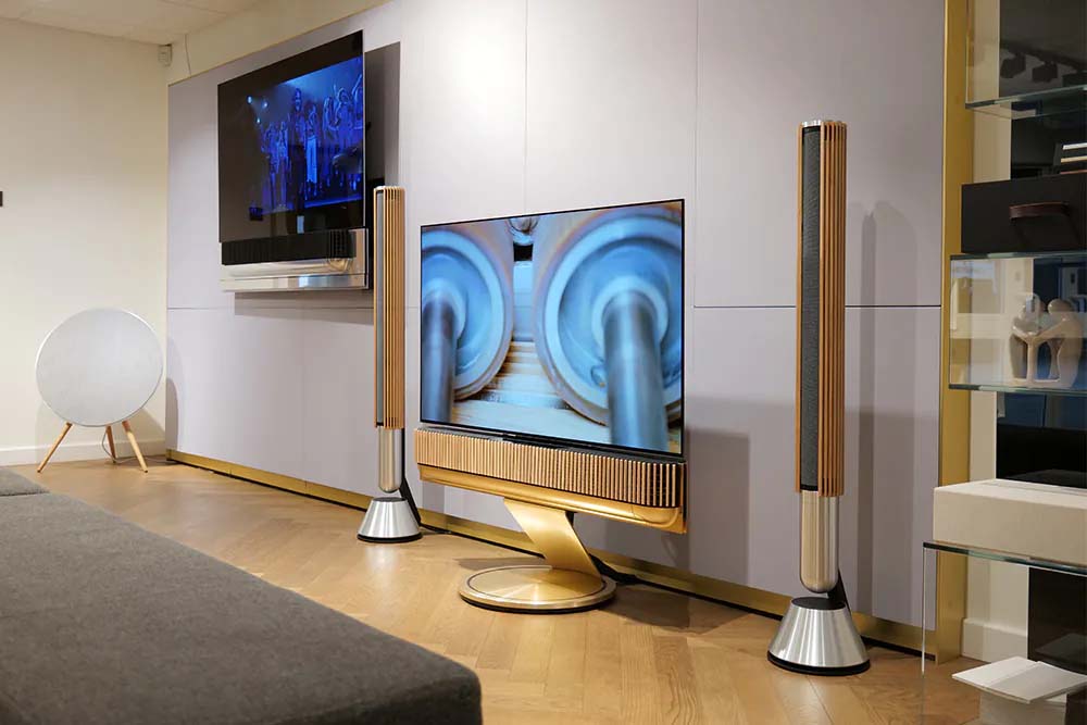 Modern living room with sleek design. A large TV on a golden stand is flanked by tall speakers. A second TV is mounted on the wall, all set on a wooden floor.