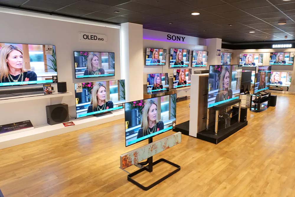 Electronics store displaying multiple flat-screen TVs on light wood flooring. Each TV shows the same image of a woman talking, creating a modern, tech-focused atmosphere.