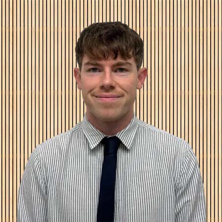 A smiling person with short brown hair wears a striped shirt and dark tie, standing against a background of vertical wooden slats. The tone is formal and friendly.