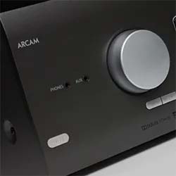 Close-up of a black Arcam audio component with a large silver knob on the front. Buttons are labeled for phono, aux, and other inputs, giving a sleek, modern feel.