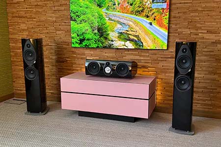 A modern home entertainment setup features a wall-mounted TV displaying a scenic landscape. Below is a pink console with a central speaker, flanked by two tall black speakers, set against a textured wooden wall.