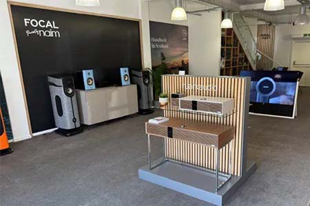 Modern audio showroom with sleek speakers and a wooden display stand in a spacious, well-lit room. Branding on black wall; minimalist design.
