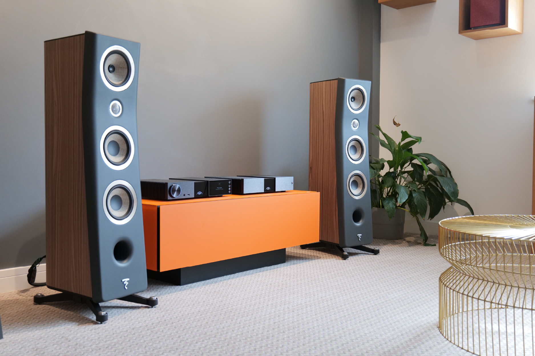 Modern living room with large wooden speakers flanking an orange console holding audio equipment. A green plant and gold wire table add warmth.