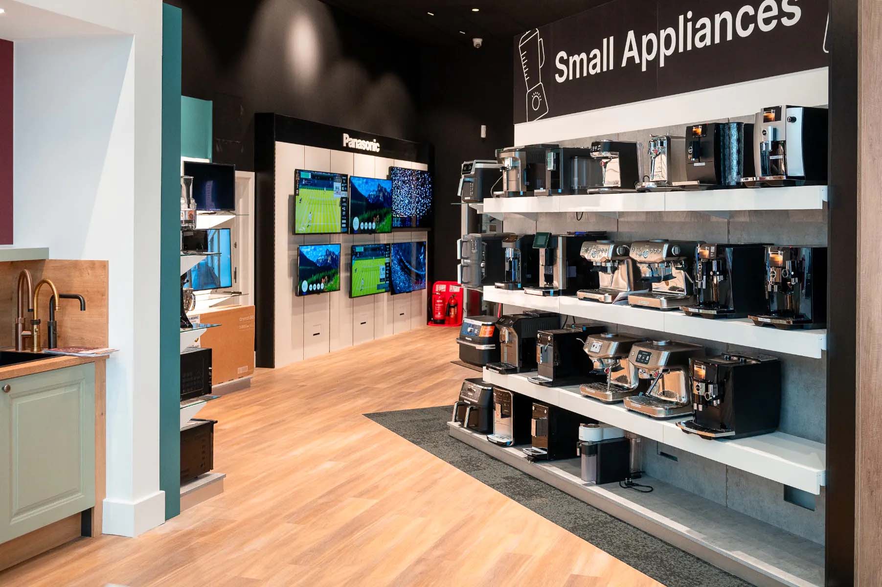 Store interior with wooden flooring. Shelves display numerous coffee machines labeled "Small Appliances." Wall-mounted TVs show various images.