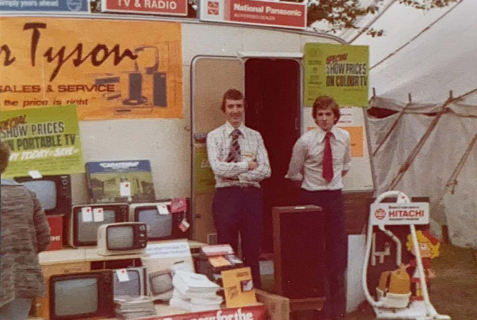 Two men in dress shirts and ties stand in front of a vintage electronics stall, showcasing various retro televisions. A large orange sign reads "Tyson Sales & Service," and the display includes posters promoting deals. The scene feels nostalgic, capturing a 1970s trade show vibe.