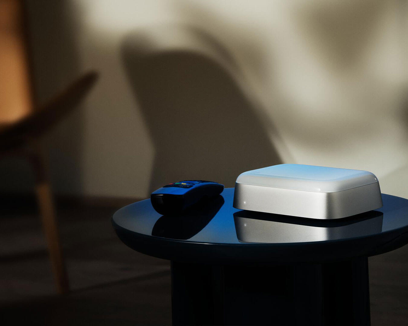 A sleek, reflective black table holds a silver electronic device and a remote control. Soft, muted lighting creates a calm and minimalist ambiance.