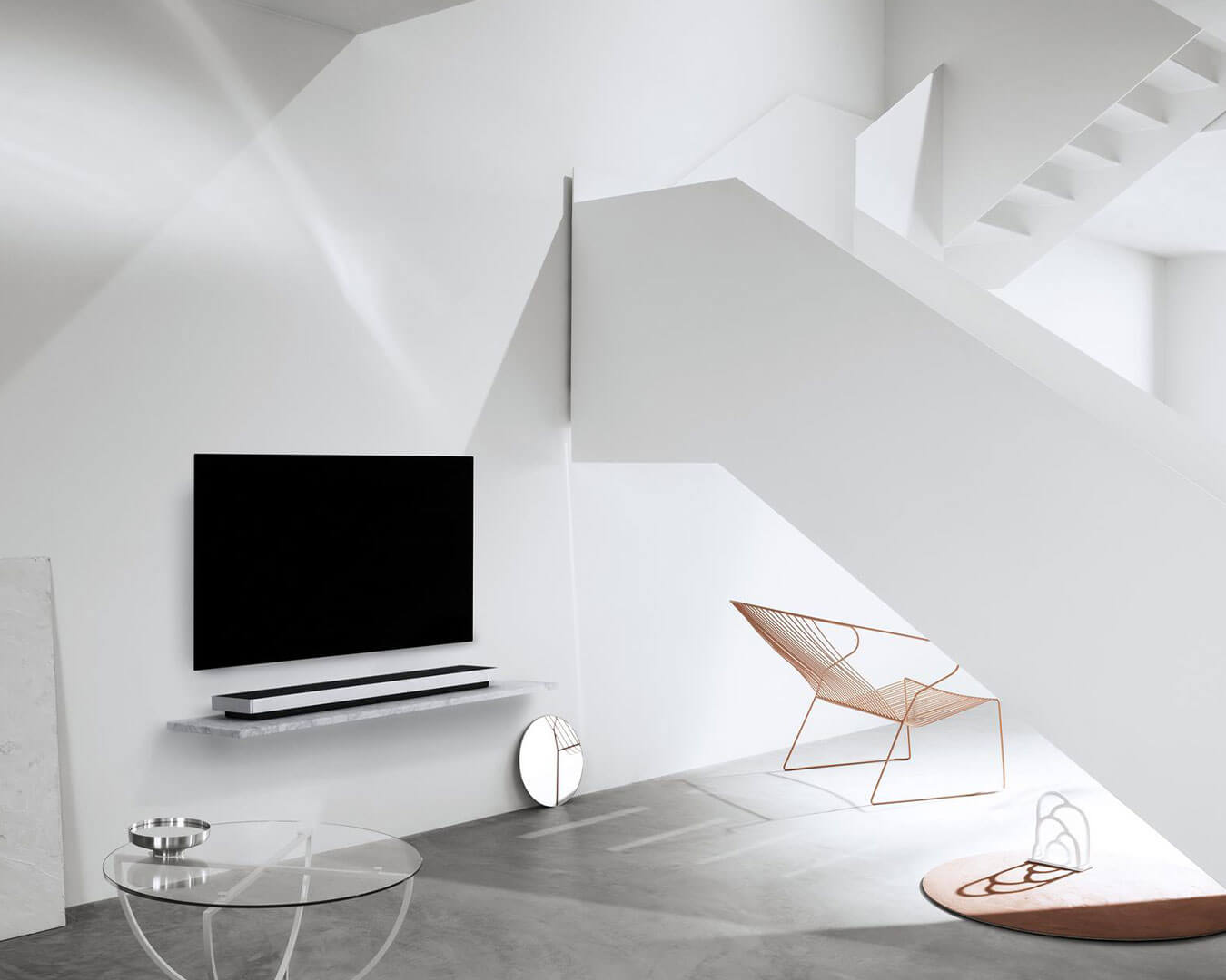 A minimalist white living room with a wall-mounted TV, a sleek glass table, and a modern wire chair. Sunlight softly highlights geometric shadows.