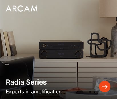 Arcam promotional banner for the Radia Series high-fidelity audio system with the headline "Radia Series" and subtext "Experts in amplification". The banner showcases two stacked, sleek black audio components—an integrated amplifier and a matching CD player—arranged elegantly on a modern cream-colored slatted sideboard cabinet. The lifestyle setting includes a minimalist room decor with books to the left, an abstract black wire loop sculpture, a stone base table lamp to the right, and the prominent white Arcam brand logo in the top-left corner. An orange circular action button with a white arrow is located at the bottom-right corner.