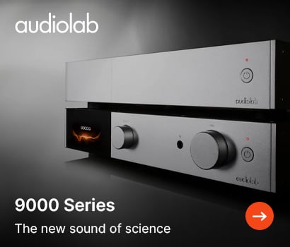 Audiolab promotional banner for the 9000 Series hi-fi system featuring the headline "9000 Series" and the subtext "The new sound of science". The banner shows a close-up of two stacked, premium silver metallic audio components angled toward the left. The bottom unit features a digital display screen on the left showing orange flame-like graphic frequencies alongside physical control dials and a power button, while the top component has a clean minimalist faceplate with a single power button. The prominent white Audiolab brand logo is visible at the top left, and an orange circular action button with a white arrow is positioned in the bottom right corner.