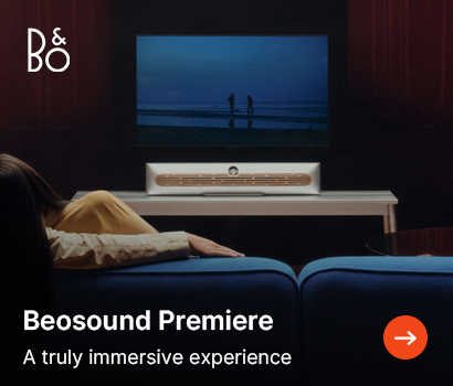 Bang & Olufsen promotional banner for the Beosound Theatre soundbar featuring the headline "Beosound Premiere" and subtext "A truly immersive experience". The image shows a dimly lit living room setting from behind a blue sofa, where a person is lying down watching a television screen that displays two figures on a beach at dusk. A premium silver and wood Beosound Theatre soundbar sits on a light-colored media console beneath the TV. The white B&O logo is positioned at the top left corner, and a bright orange circular action button with a white right-pointing arrow is at the bottom right.