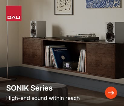 DALI promotional banner for the SONIK Series loudspeakers featuring the headline "SONIK Series" and subtext "High-end sound within reach". The banner shows a lifestyle interior with a pair of compact grey bookshelf speakers placed on top of a floating dark wood media console cabinet. The central open shelf of the console holds vinyl records and a compact black integrated amplifier, while a turntable rests on top. The prominent red square DALI logo is located in the top-left corner, and an orange circular action button with a white right-pointing arrow sits in the bottom-right corner.