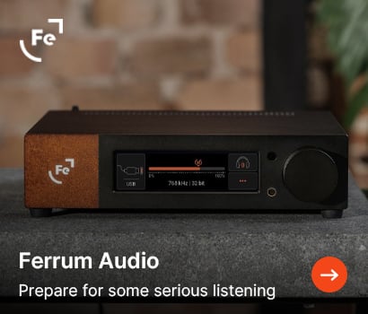 Ferrum Audio promotional banner showcasing a premium high-end DAC and headphone amplifier. The dark metallic device features a distinctive textured rust-colored accent patch on the left side with a glowing white "Fe" logo. The central digital screen displays system information including a volume slider bar, "USB" input selection, and "768 kHz | 32 bit" audio resolution readout. The unit rests on a grey stone surface against a blurred background of a brick wall and a houseplant. The top left shows the white Ferrum Audio brand symbol, while the bottom highlights the text "Ferrum Audio" and "Prepare for some serious listening" next to an orange circular action button with a white arrow.