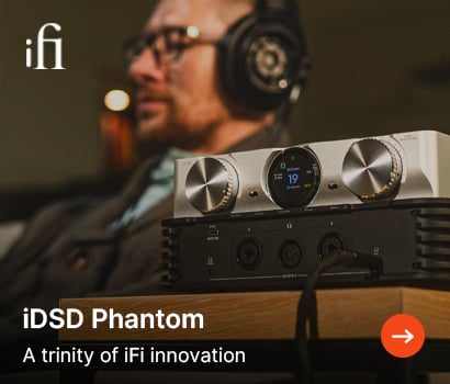 iFi promotional banner for the iDSD Phantom. The foreground shows a close-up of the silver and black premium headphone amplifier and DAC unit sitting on a wooden desk. In the blurred background, a man wearing large over-ear open-back headphones listening intently. The white iFi brand logo sits at the top left corner, with text at the bottom left reading "iDSD Phantom" and "A trinity of iFi innovation" next to a circular orange action button with a white right-pointing arrow.