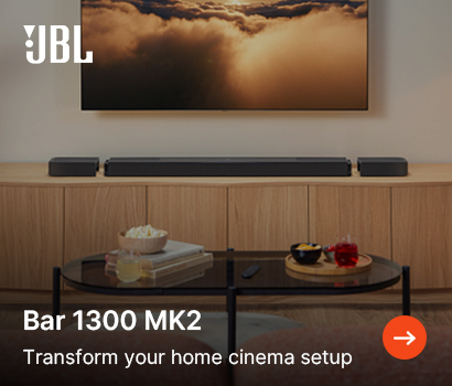 JBL promotional banner for the Bar 1300 MK2 home cinema system featuring the headline "Bar 1300 MK2" and subtext "Transform your home cinema setup". The image shows a cozy living room home theater arrangement where the long black soundbar sits on a light wood credenza beneath a wall-mounted TV displaying golden, sunlit clouds. The system's detachable wireless surround speakers are positioned symmetrically on either side of the main bar. In the foreground, an oval black glass coffee table holds snacks and drinks. The white JBL logo is placed at the top-left corner, and a bright orange circular action button with a white right-pointing arrow sits at the bottom right.