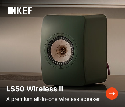 KEF promotional banner for the LS50 Wireless II loudspeaker featuring the headline "LS50 Wireless II" and subtext "A premium all-in-one wireless speaker". The banner features a close-up view of a single forest green bookshelf speaker with a distinctive copper-colored Uni-Q driver array. The speaker sits on a sleek, reflective countertop surface inside a modern kitchen or living area. The white KEF brand logo is located at the top-left corner, and an orange circular action button with a white right-pointing arrow is positioned in the bottom-right corner.