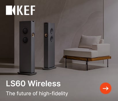 KEF promotional banner for the LS60 Wireless loudspeakers featuring the headline "LS60 Wireless" and subtext "The future of high-fidelity". The banner shows a modern, minimalist room setting with a pair of sleek, slim grey floorstanding speakers standing on a polished floor. A contemporary off-white armchair with a wooden base and black metal legs is positioned to the right against a textured neutral wall. The prominent white KEF brand logo sits at the top left corner, and an orange circular action button with a white right-pointing arrow is located in the bottom right corner.