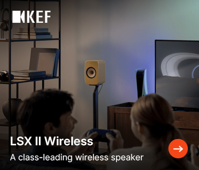 KEF promotional banner for the LSX II Wireless loudspeaker system featuring the headline "LSX II Wireless" and subtext "A class-leading wireless speaker". The image shows a man and a woman sitting on a couch in a living room, playing a video game with a blue controller while facing a television screen. In the background, a compact cream or tan bookshelf speaker is mounted on a sleek black floor stand next to a PlayStation 5 console and a tall shelving unit filled with books. The white KEF logo is placed in the top left corner, and a bright orange circular button with a white right-pointing arrow is at the bottom right.
