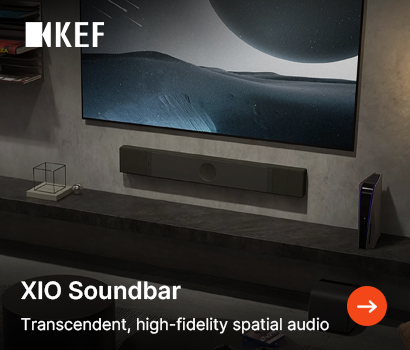 KEF promotional banner for the XIO Soundbar featuring the headline "XIO Soundbar" and subtext "Transcendent, high-fidelity spatial audio". The banner shows a modern, minimalist home theater setup with a slim black soundbar mounted to a concrete-textured wall directly underneath a large flat-screen television. The television display shows a black-and-white cinematic cosmic landscape image. A dark floating shelf beneath the soundbar holds a PlayStation 5 console and a minimalist wire-frame decorative cube. The white KEF logo is located in the top-left corner, and an orange circular action button with a white right-pointing arrow sits in the bottom-right corner.