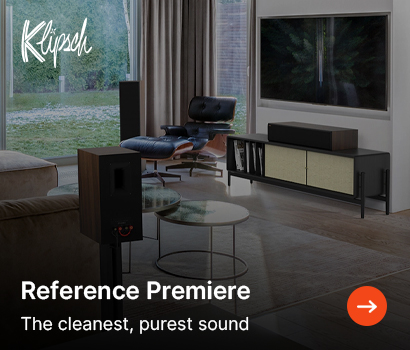 Klipsch promotional banner for the Reference Premiere speaker series featuring the headline "Reference Premiere" and subtext "The cleanest, purest sound". The banner shows a modern, stylish living room interior equipped with a full home cinema audio setup. A bookshelf speaker sits on a stand in the foreground, a center channel speaker rests on a dark media console below a large wall-mounted television, and a floorstanding tower speaker stands near a classic leather lounge chair and ottoman in the background. The white script Klipsch logo is located in the top left corner, and an orange circular action button with a white right-pointing arrow is positioned in the bottom right corner.