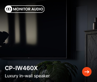 Monitor Audio promotional banner for the CP-IW460X speaker featuring the headline "CP-IW460X" and subtext "Luxury in-wall speaker". The banner features a dimly lit, moody interior setting showing a large wall-mounted flat-screen television. A flush, rectangular white speaker grille is seamlessly integrated into the wall to the right of the television screen. The scene includes a dark wooden cabinet on the right and a houseplant positioned in the lower foreground. The white Monitor Audio logo sits at the top left corner, and an orange circular action button with a white right-pointing arrow is located in the bottom right corner.