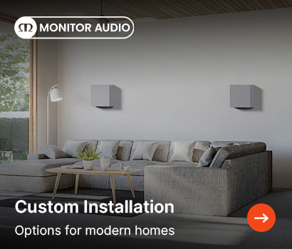 Monitor Audio promotional banner for custom installation audio solutions featuring the headline "Custom Installation" and subtext "Options for modern homes". The banner showcases a bright, modern living room interior with a large grey L-shaped sectional sofa arranged around a light wood coffee table. Two square, light grey on-wall speakers are mounted symmetrically on the back wall above the sofa cushions. The background features a tall white floor lamp to the left near a large glass door looking out onto greenery, with the white Monitor Audio logo in the top left corner and an orange circular action button with a white right-pointing arrow in the bottom right corner.