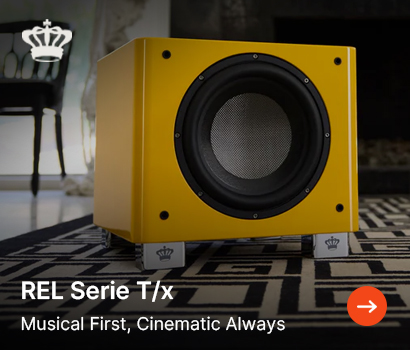 REL Acoustics promotional banner for the Serie T/x subwoofer featuring the headline "REL Serie T/x" and subtext "Musical First, Cinematic Always". The banner showcases a bright yellow cube subwoofer resting on silver feet bearing the brand's crown logo. The device is positioned on a black and white geometric patterned rug within a dimly lit living room setting, with a classic chair visible in the background. A large white crown logo is placed in the top left corner, and an orange circular action button with a white right-pointing arrow is located in the bottom right corner.