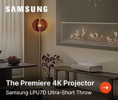 Samsung promotional banner for the Premiere 4K Projector, specifically identifying the Samsung LPU7D Ultra-Short Throw model. The image showcases a modern home interior where the white ultra-short-throw projector sits on a wooden bench, projecting an image onto the wall above an integrated linear fireplace with small flames. The cozy room setting includes a glowing spherical wooden floor lamp, a small houseplant, a wall clock, and a dining table vignette to the left. The prominent white Samsung logo is at the top left, and a bright orange circular action button with a white arrow sits at the bottom right.