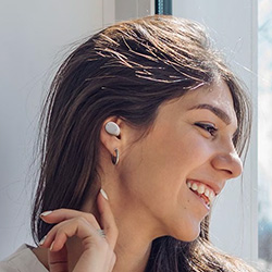 Smiling woman with long brown hair wears a white earbud, looking out a bright window peacefully. Sunlight highlights her relaxed expression.