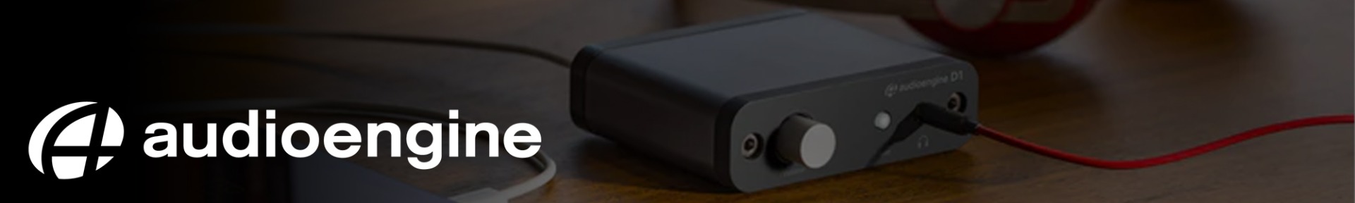 A compact black audio interface with a volume knob and connected red cable sits on a wooden surface. "Audioengine" logo on the left. Modern and sleek design.
