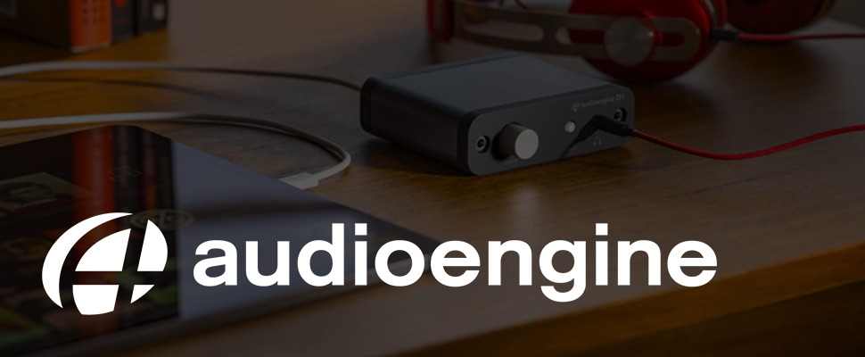 A compact black audio interface with a volume knob and connected red cable sits on a wooden surface. "Audioengine" logo on the left. Modern and sleek design.