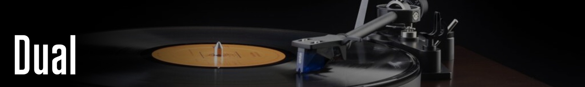 Turntable playing a vinyl record with a yellow label, set on a dark wooden surface. The word "Dual" appears in white on the left side.