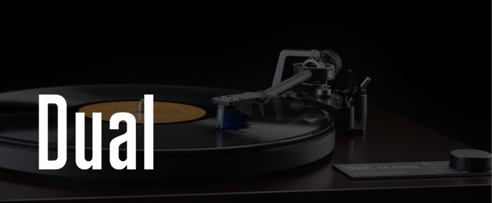 Turntable playing a vinyl record with a yellow label, set on a dark wooden surface. The word "Dual" appears in white on the left side.