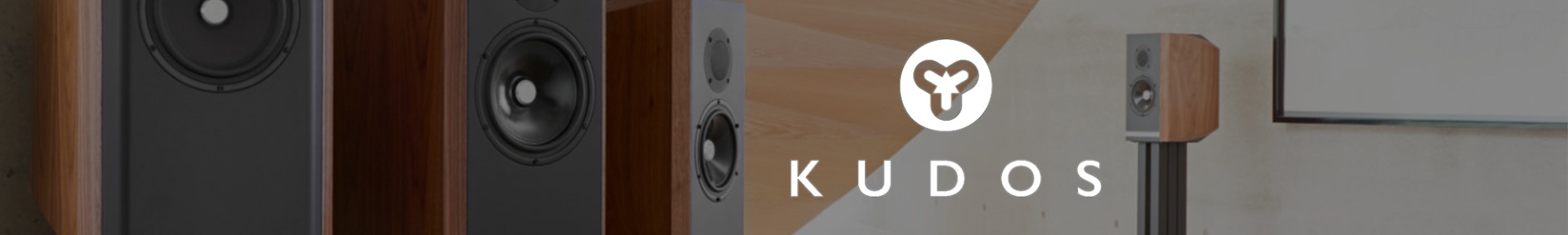 Close-up of stylish wooden speakers with black fronts, featuring the "Kudos" logo in white. The setting is modern, conveying a sleek, sophisticated tone.