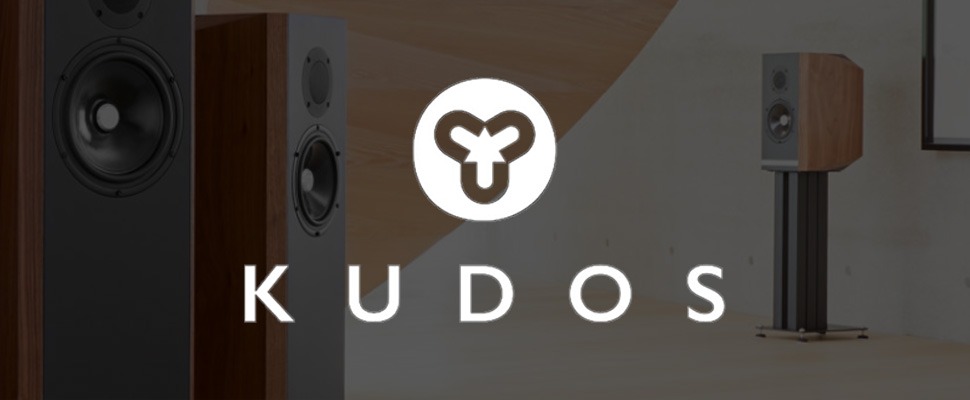 Close-up of stylish wooden speakers with black fronts, featuring the "Kudos" logo in white. The setting is modern, conveying a sleek, sophisticated tone.