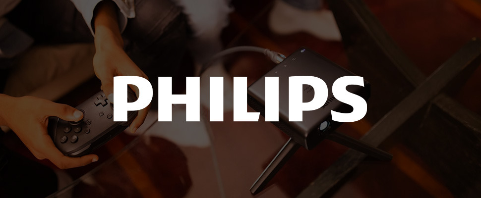 A Philips mini projector sits on a table, projecting light. A hand holds a game controller nearby. The scene conveys technology and entertainment integration.