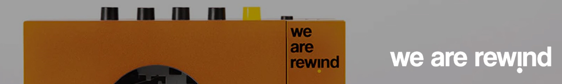 Close-up of an orange retro-style cassette player with black and yellow buttons on top. The text "we are rewind" is printed in black on the device. The background is grey.