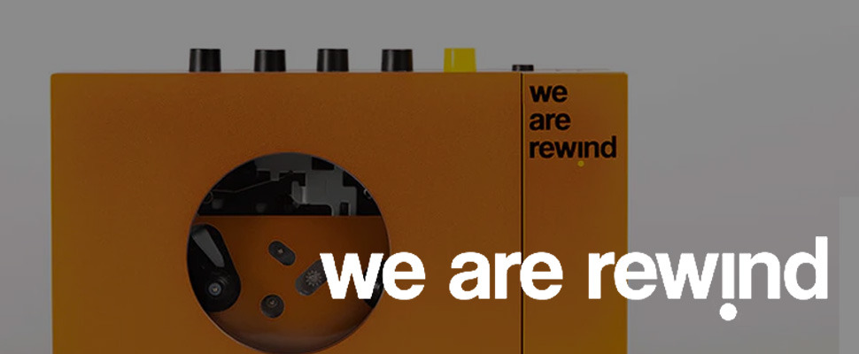 Close-up of an orange retro-style cassette player with black and yellow buttons on top. The text "we are rewind" is printed in black on the device. The background is grey.