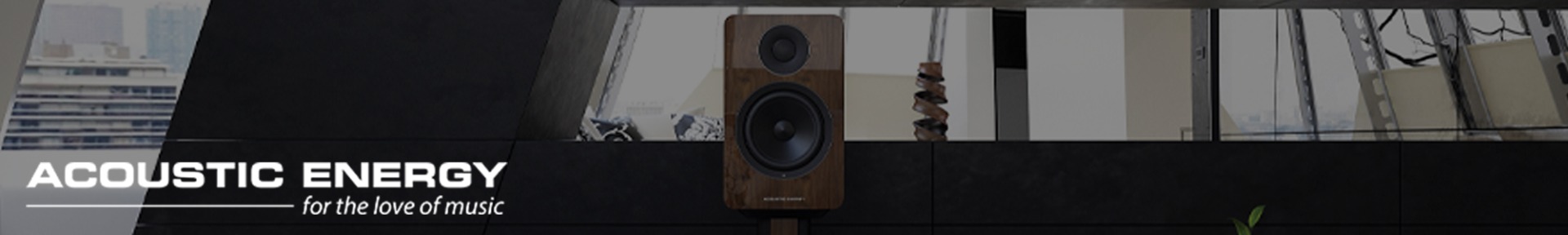 A sleek wooden speaker on a stand is centred in a modern room with large windows. The text "ACOUSTIC ENERGY: for the love of music" is on the left.