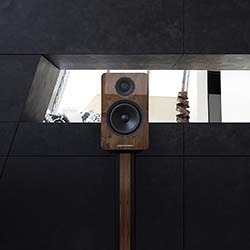A wooden speaker on a stand is centred in a modern, dimly lit room with dark, geometric walls and a small, bright window above. The mood is sleek and minimalist.