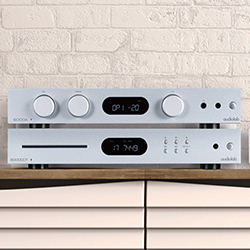 Silver audio system with two stacked units: one with large knobs and a digital display, the other a CD player, set against a brick wall backdrop.