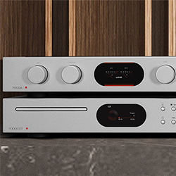 Sleek silver audio components on a marble surface against a wood backdrop. Top unit has knobs and a digital screen; bottom has buttons and a CD slot. Modern and elegant.
