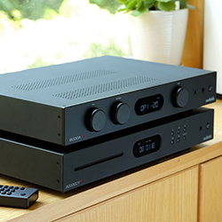 A sleek black stereo receiver and CD player stack on a wooden shelf, with a remote nearby. Sunlight and plants create a calm, modern atmosphere.