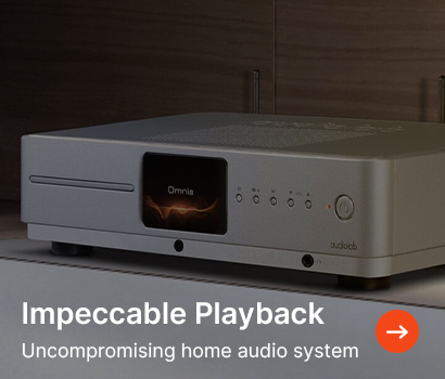 A sleek silver audio system on a shelf, displaying "Omnia" on its screen. Caption reads "Impeccable Playback" with an arrow icon for more info.