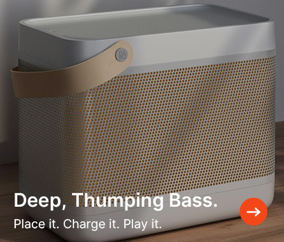 Compact speaker with modern design sits on a wooden surface. Features a leather strap and perforated speaker grill. Text reads "Deep, Thumping Bass."