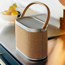 A stylish, portable speaker with a woven texture and leather handle sits on a modern kitchen counter. A bowl of lemons is in the soft-focus background.