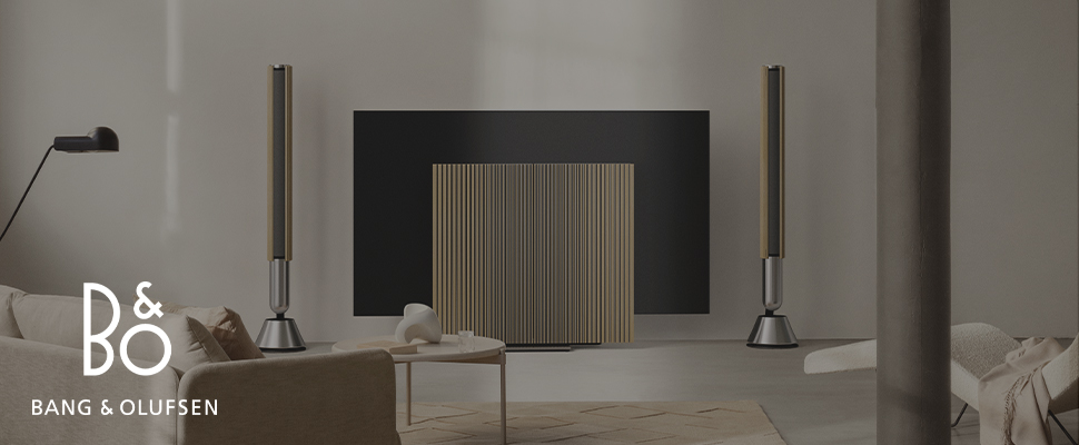 Modern living room with minimalist decor. Features tall speakers, a sleek TV, stylish lighting, and lush plants. Bang & Olufsen logo displayed.