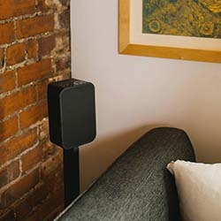 A modern speaker on a stand next to a grey couch with a white pillow in a cosy room. The wall has exposed brick and a framed abstract artwork.