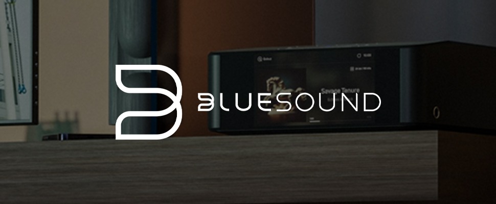 A sleek audio player displays "Savage Tenure" on a screen, surrounded by speakers and wood accents. Bluesound logo is on the left, adding a modern touch.