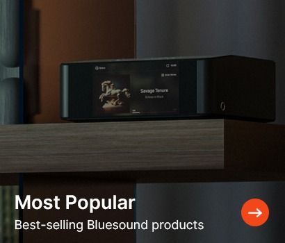 A sleek black Bluesound audio device sits on a wooden shelf displaying a song on its screen. Text reads "Most Popular, Best-selling Bluesound products" with an arrow icon.