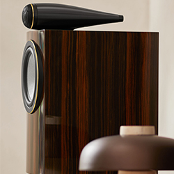 Wooden high-end speaker with sleek black top cone and reflective finish, next to blurred lamp. Modern, elegant design in a sophisticated setting.